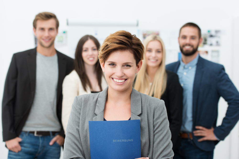 Group of job applicants with a smiling confident young woman in the foreground holding her Curriculum vitae in her hands Group of job applicants with a smiling confident young woman in the foreground holding her Curriculum vitae in her hands